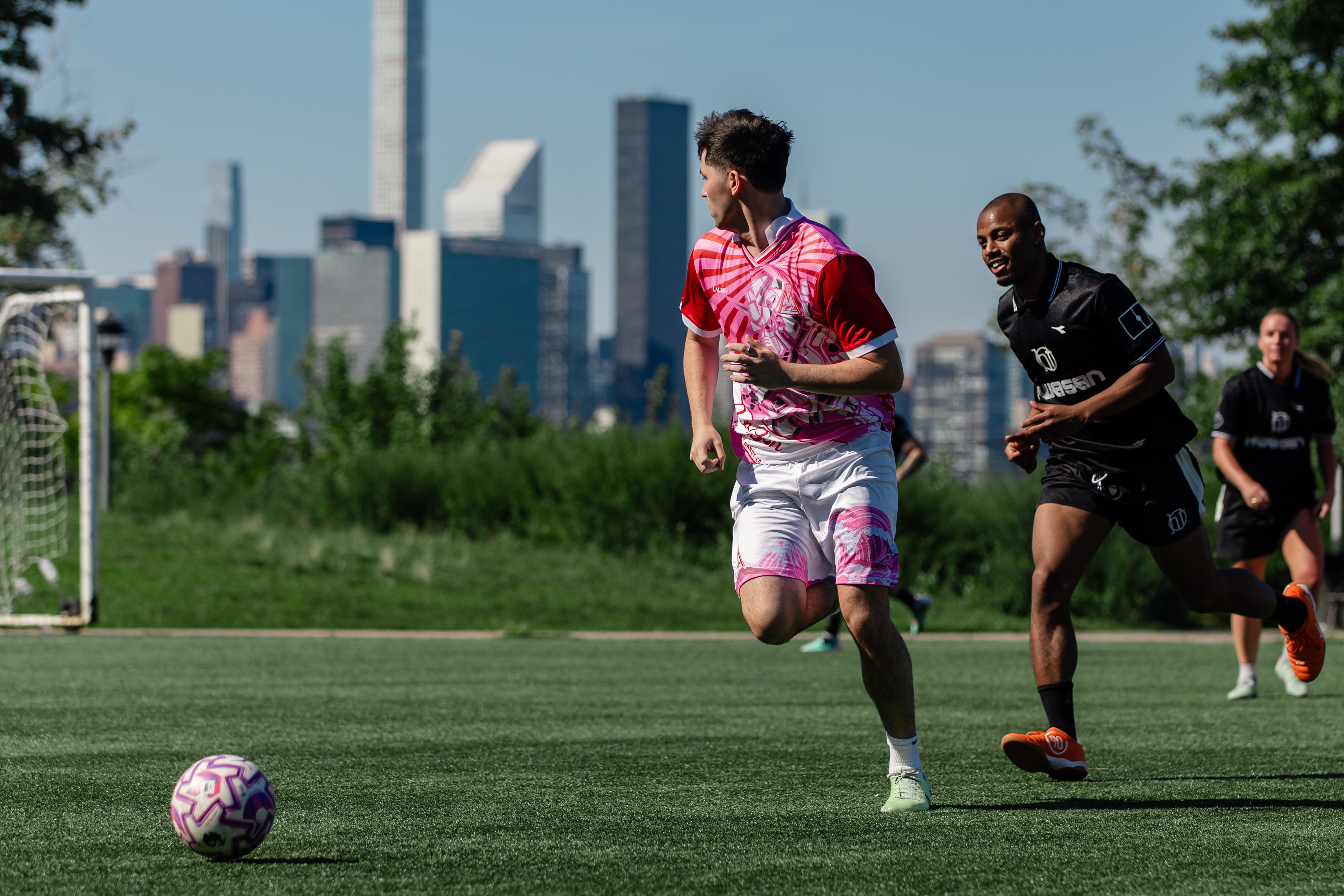 A man in a pink and white soccer jersey and shorts runs on a green field while another player in a black jersey chases him, with a city skyline and trees in the background.